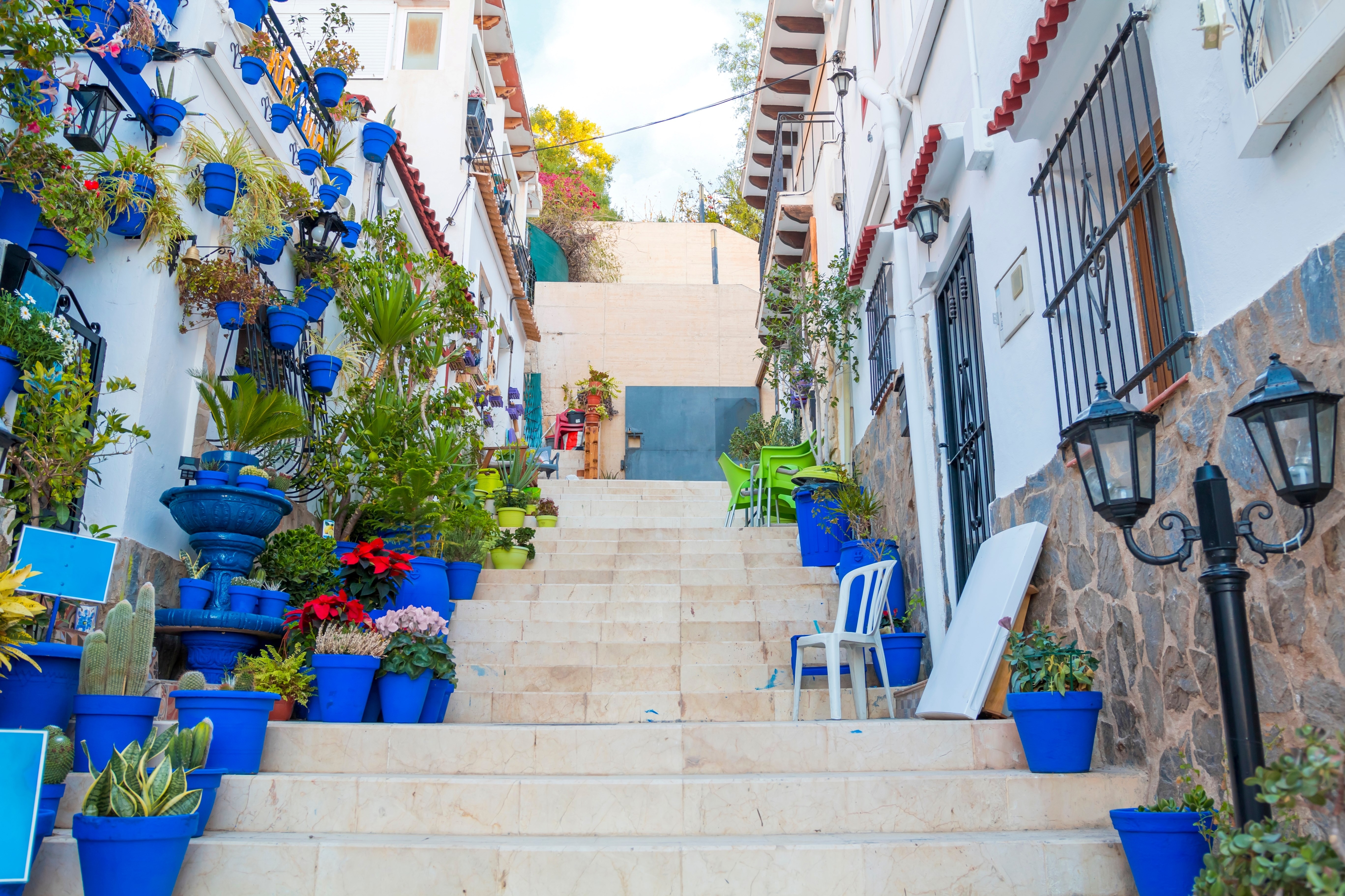 Narrow street with steps, white houses and blue potted plants in ancient neighborhood Santa Cruz in Alicante old town on hillside. Costa Blanca on Mediterranean sea coast, Spain