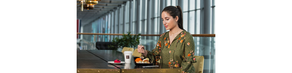 A woman in a green top at a table eating with chopsticks, a coffee mug and a pastry in front of her A woman in a green top at a table eating with chopsticks, a coffee mug and a pastry in front of her